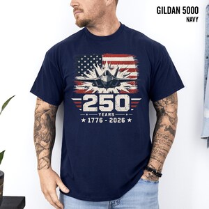 May include: Navy blue Gildan 5000 t-shirt featuring a graphic of a fighter jet in front of a distressed American flag. The design includes the text "250 YEARS 1776-2026".