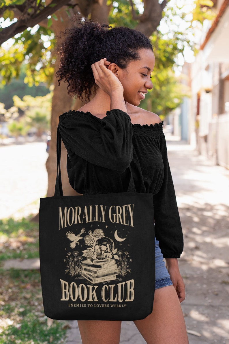 May include: A black tote bag with a white design that says "Morally Grey Book Club" with a skull, roses, and a bird. The text below the design says "Enemies to Lovers Weekly."