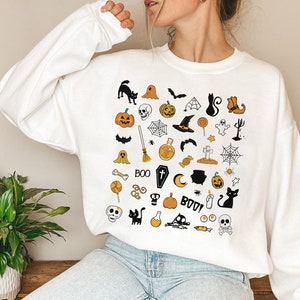 May include: White sweatshirt with a black and orange Halloween themed print. The print features a variety of Halloween icons, including pumpkins, ghosts, bats, witches hats, and candy corn. The text "BOO!" is also included in the print.