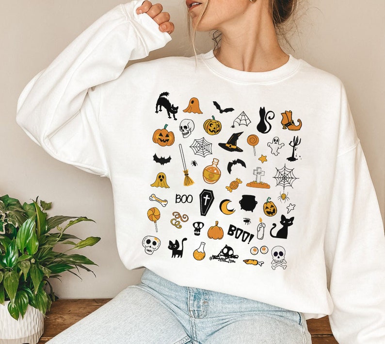 May include: White sweatshirt with a black and orange Halloween themed print. The print features a variety of Halloween icons, including pumpkins, ghosts, bats, witches hats, and candy corn. The text "BOO!" is also included in the print.