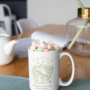 May include: White ceramic mug with a green illustration of a mushroom, moon phases, and butterflies. The text "We shall meet in the place where there is no darkness." is printed on the mug. The mug is filled with pastel-colored marshmallows.