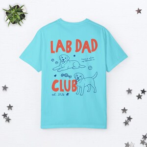 Puede incluir: Una camiseta azul claro con el texto rojo LAB DAD CLUB. El dise&ntilde;o incluye dos ilustraciones de l&iacute;nea de perros Labrador, huellas, una pelota, un juguete y el texto home is where my labrador is y est. 2026 en tinta azul oscuro.