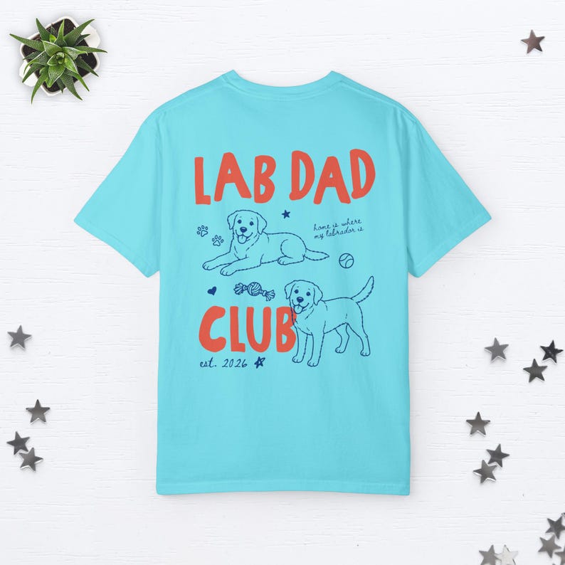 Puede incluir: Una camiseta azul claro con el texto rojo LAB DAD CLUB. El dise&ntilde;o incluye dos ilustraciones de l&iacute;nea de perros Labrador, huellas, una pelota, un juguete y el texto home is where my labrador is y est. 2026 en tinta azul oscuro.