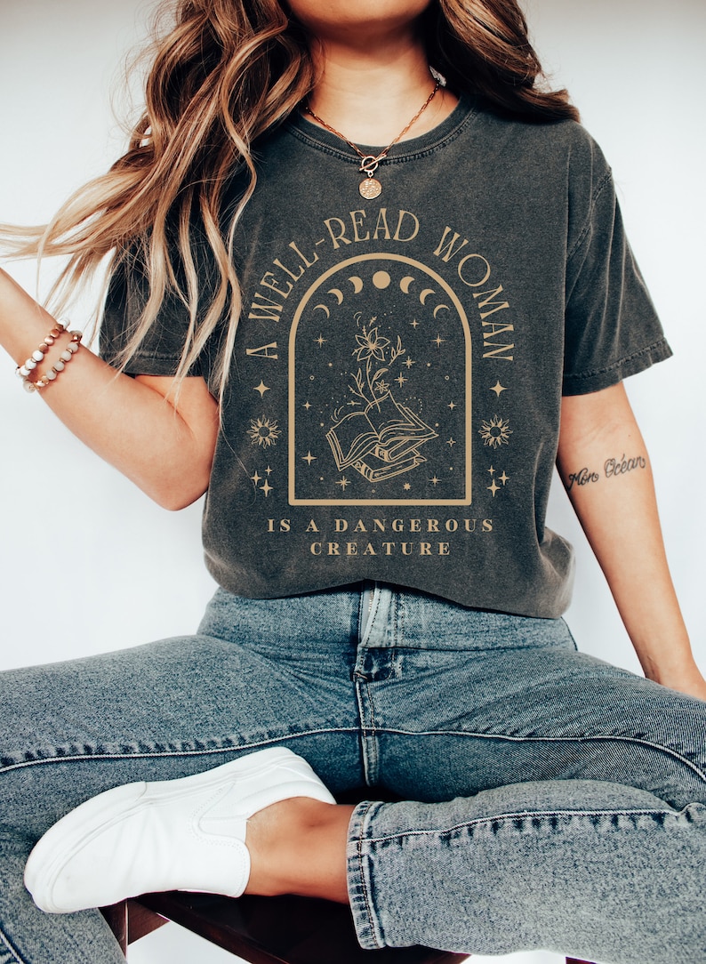 May include: A dark gray t-shirt with a graphic design featuring a book, flowers, and stars. The text on the shirt reads "A well-read woman is a dangerous creature."