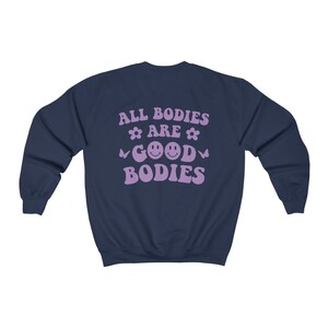 May include: Navy blue sweatshirt with lavender text reading "ALL BODIES ARE GOOD BODIES". The design includes flowers, butterflies, and smiley faces. A casual, comfortable style of clothing.