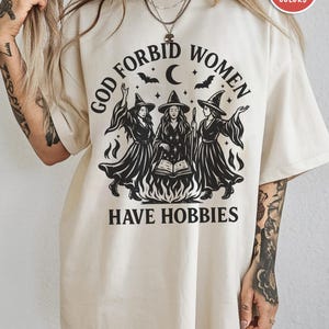 May include: An ivory t-shirt featuring a black graphic print of three witches standing around a campfire with an open book. The text reads "GOD FORBID WOMEN HAVE HOBBIES" in a curved font above the witches, with bats and a crescent moon above.