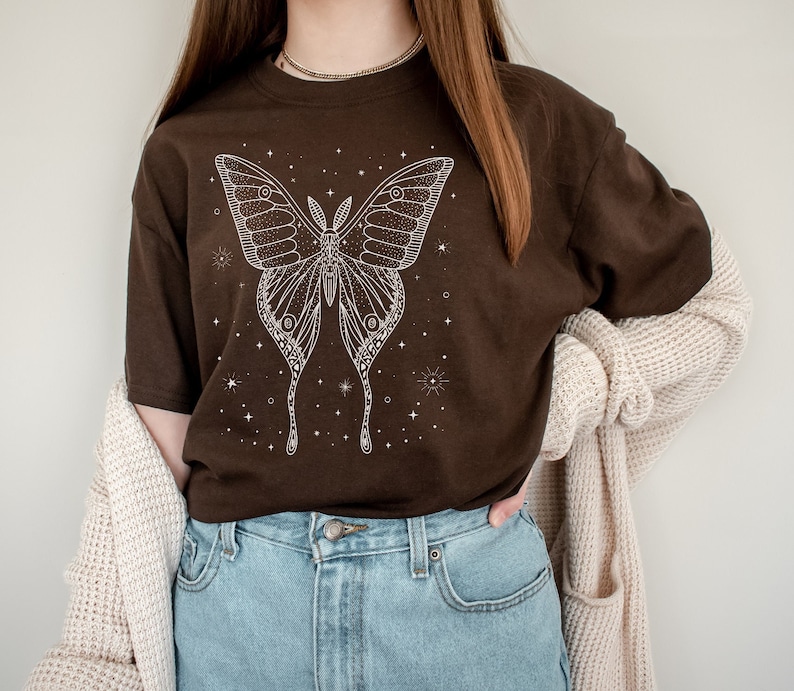 May include: A brown t-shirt with a white outline of a moth with stars and a celestial theme. The shirt is being worn by a person with a brown cardigan.