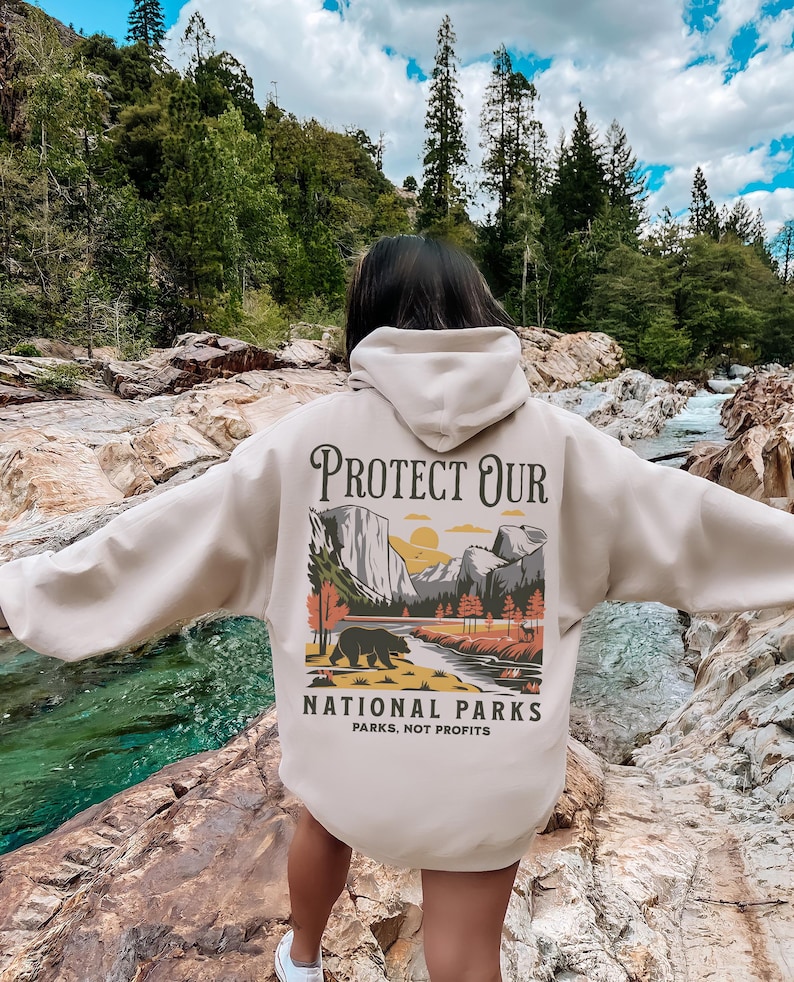 May include: A beige hoodie with a graphic on the back that reads "PROTECT OUR NATIONAL PARKS" with an illustration of a bear, mountains, and trees. The text "PARKS, NOT PROFITS" is below the illustration. The hoodie is being worn outdoors.