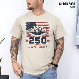 May include: Sand-colored Gildan 5000 t-shirt featuring a graphic of an American flag, a jet, and the text "250 YEARS 1776-2026". The design is in red, white, and blue.
