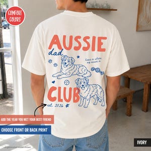 May include: An ivory t-shirt featuring a back graphic with the text "AUSSIE DAD CLUB" in large coral letters. The design includes blue line art of two Australian Shepherd dogs, paw prints, a ball, a toy, stars, and the text "home is where my aussie is" and "est. 2026".