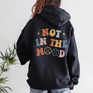 Pu&ograve; includere: Felpa con cappuccio nera con il testo "NOT IN THE MOOD" in colori arcobaleno sul retro.