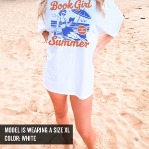 K&ouml;nnte beinhalten: Wei&szlig;es T-Shirt mit einer blauen und orangenen Grafik einer Frau, die ein Buch am Strand liest, mit dem Text "Book Girl Summer" und "Chapters and Sunshine" in einem Kreis.