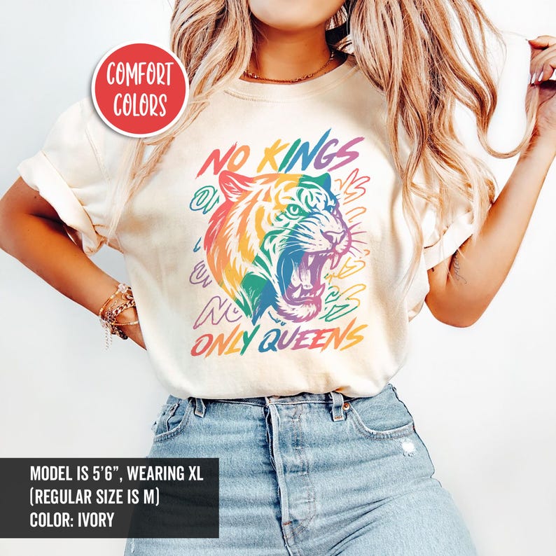 May include: Ivory-colored t-shirt with a rainbow tiger graphic and the text "NO KINGS ONLY QUEENS." The design includes the words "COMFORT COLORS." The shirt is a regular fit.
