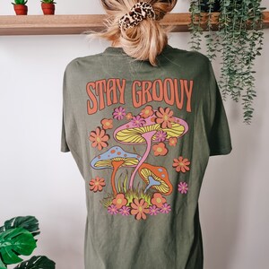 May include: Green t-shirt with a retro graphic of mushrooms and flowers. The text "Stay Groovy" is printed in orange on the shirt.