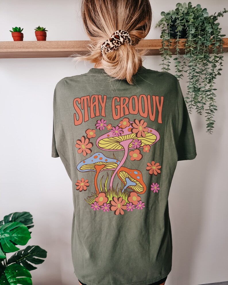 May include: Green t-shirt with a retro graphic of mushrooms and flowers. The text "Stay Groovy" is printed in orange on the shirt.