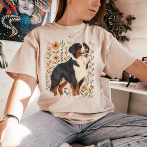 Pu&ograve; includere: Una maglietta beige con un design floreale che presenta un cane da montagna bernese. Il cane &egrave; marrone, nero e bianco con un motivo floreale dietro.
