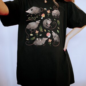 May include: Black t-shirt with a graphic print of opossums, mushrooms, and flowers. The opossums are depicted in a cartoon style with colorful accents.