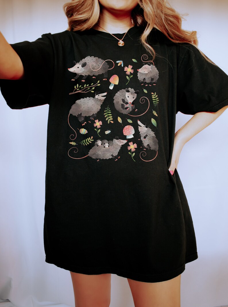 May include: Black t-shirt with a graphic print of opossums, mushrooms, and flowers. The opossums are depicted in a cartoon style with colorful accents.