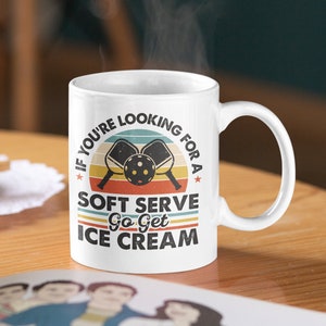 May include: White ceramic mug with a retro-style design featuring a sunset, two pickleball paddles, and the text "If you're looking for a soft serve go get ice cream".