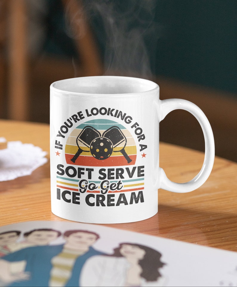 May include: White ceramic mug with a retro-style design featuring a sunset, two pickleball paddles, and the text "If you're looking for a soft serve go get ice cream".