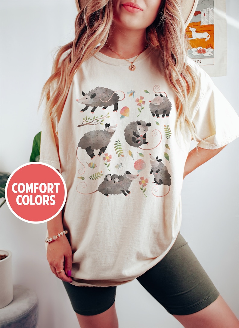 May include: A beige t-shirt with a graphic print of multiple opossums in a woodland setting. The opossums are depicted in a cartoon style with floral accents. The text "COMFORT COLORS" is visible in a red circle on the left side of the image.