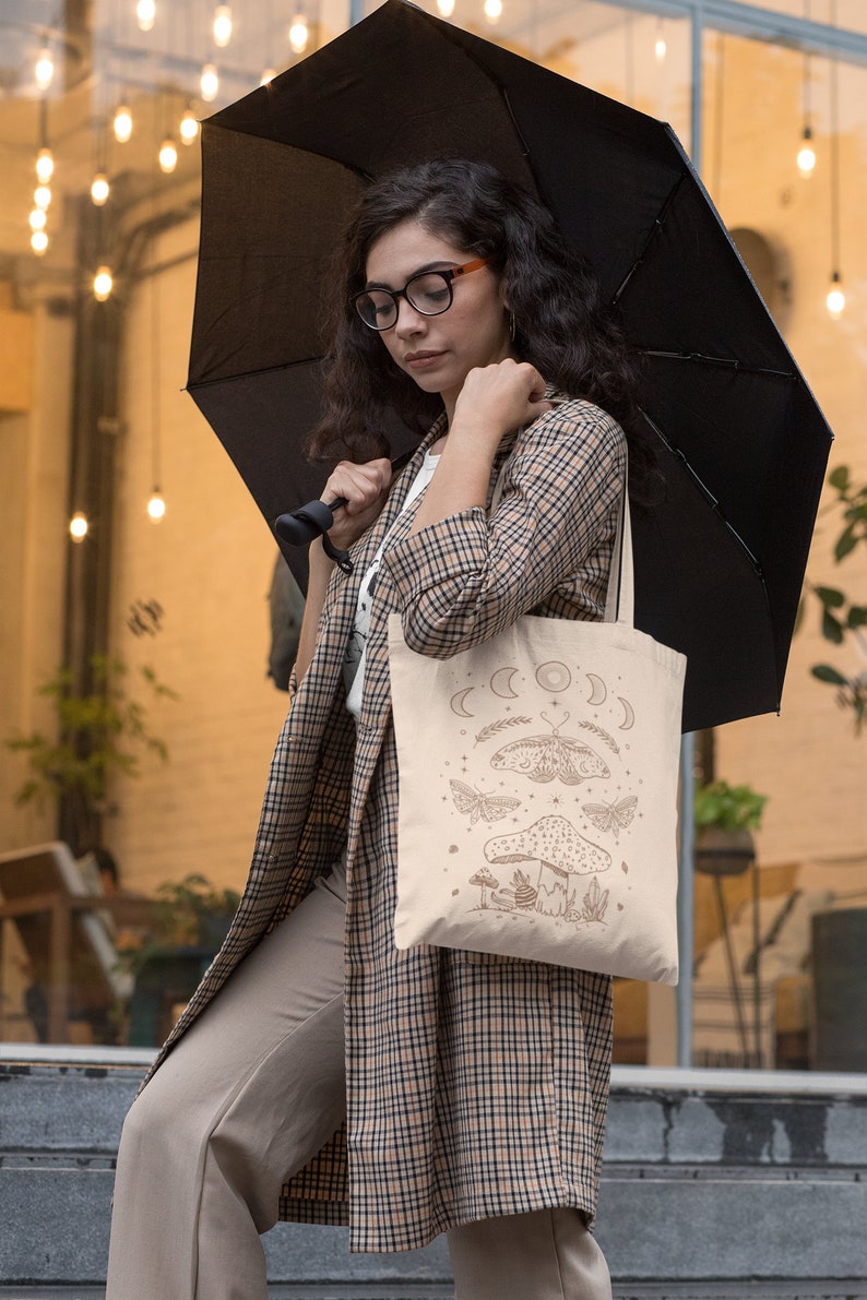 May include: A woman is holding a beige tote bag with a brown illustration of a mushroom, moths, and a crescent moon. The bag has a black strap and the woman is wearing a plaid jacket and beige pants.