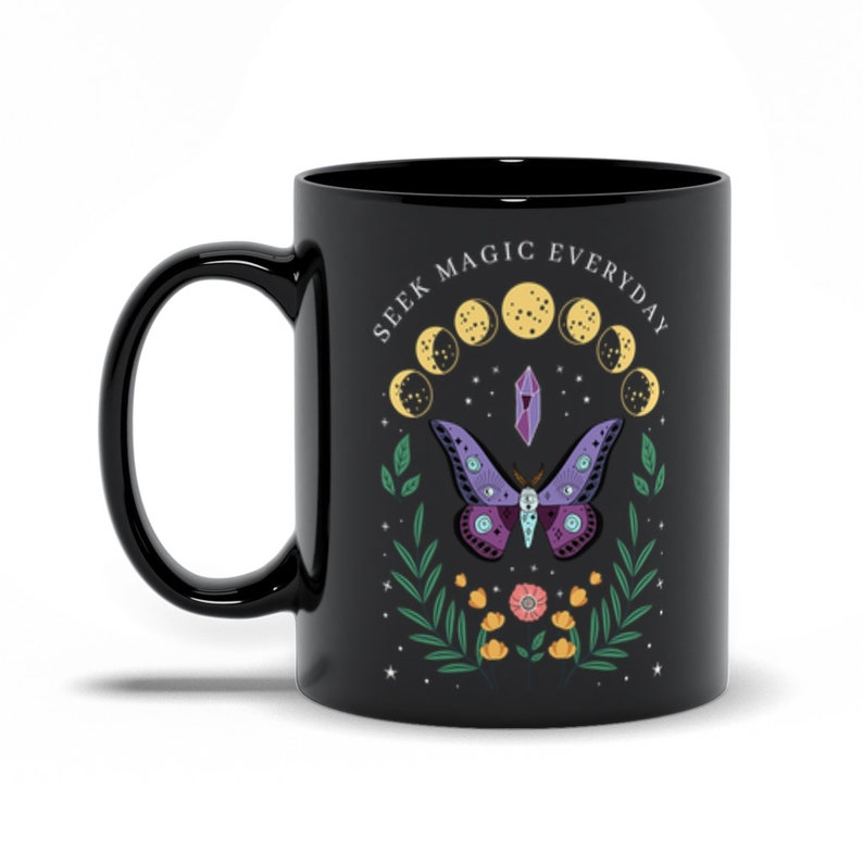May include: Black ceramic mug with a white design featuring a purple butterfly, a crystal, and a crescent moon phase cycle. The text "Seek Magic Everyday" is written in a white circular font around the moon phases.