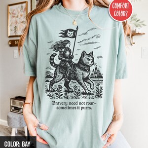 Peut inclure: Un t-shirt bleu clair avec une illustration en noir et blanc d'une femme chevauchant un chat. La femme porte une armure et tient un drapeau avec un chat dessus. Le texte "Bravery need not roar - sometimes it purrs." est imprim&eacute; sous l'illustration.