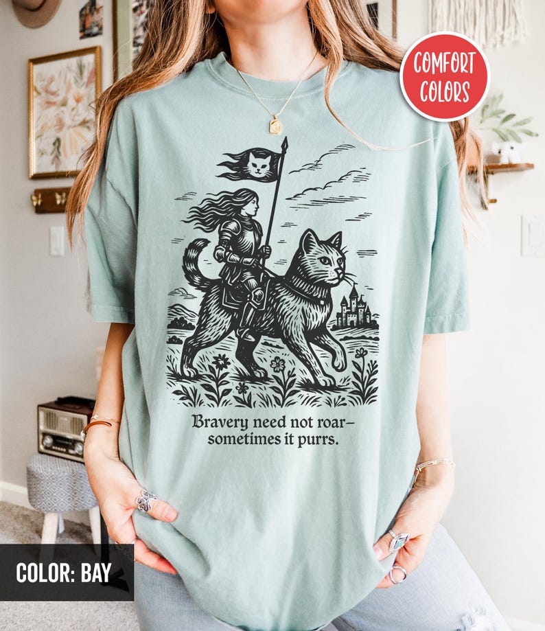 Peut inclure: Un t-shirt bleu clair avec une illustration en noir et blanc d'une femme chevauchant un chat. La femme porte une armure et tient un drapeau avec un chat dessus. Le texte "Bravery need not roar - sometimes it purrs." est imprim&eacute; sous l'illustration.