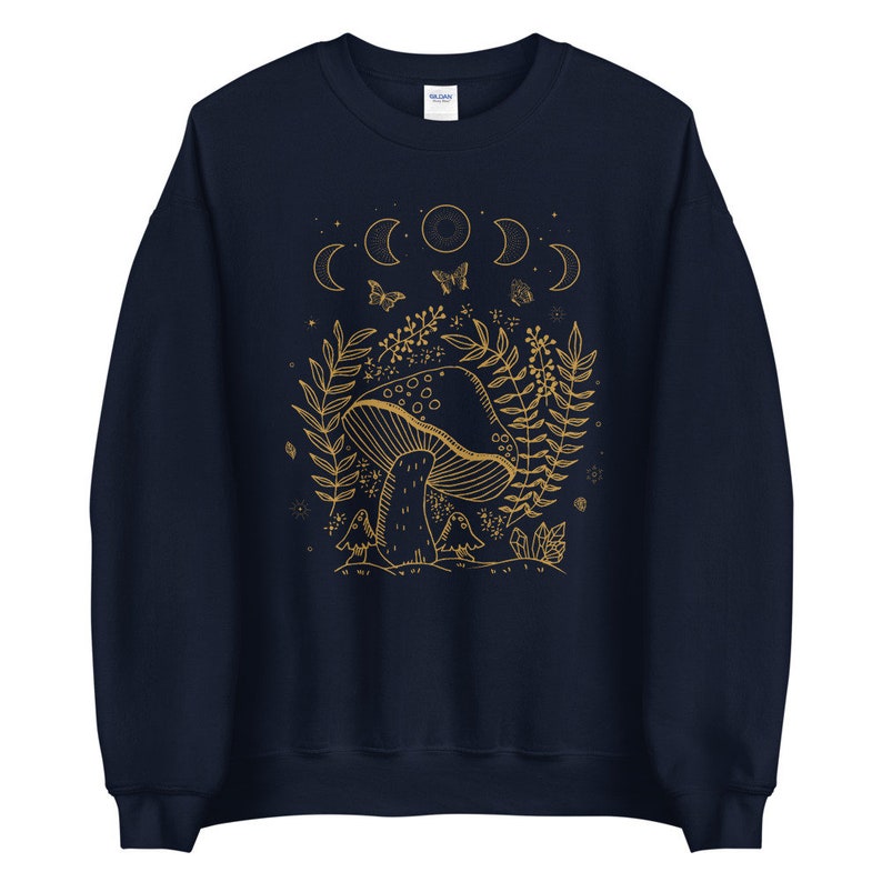 Op de afbeelding: Navyblauwe trui met een gouden omtrekillustratie van een paddenstoel, bladeren en halve manen. De tekst "Shroom Queens" is op de trui gedrukt.