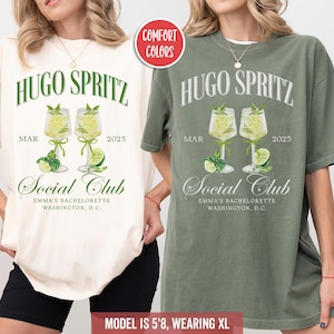 May include: Two oversized t-shirts with a green and white design. The shirts feature the text "Hugo Spritz" and "Social Club" with a graphic of two glasses of a cocktail. The shirts are available in ivory and moss.