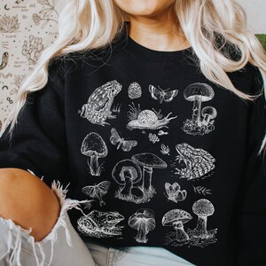 以下が含まれることがあります： 白いグラフィックプリントが施された黒いスウェットシャツ。様々なキノコ、カエル、蝶、その他の森の生き物が描かれています。