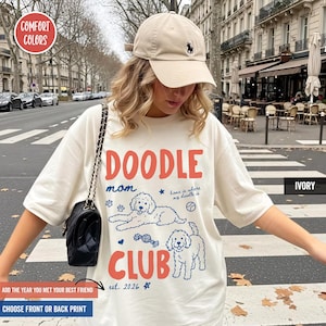 以下が含まれることがあります： アイボリー色のTシャツで、赤い文字のDOODLE CLUBと、2匹のドゥードル犬、ボール、おもちゃの青い線画が描かれています。シャツにはmom、home is where my doodle is、est. 2026という文字が含まれています。ベージュの野球帽も着用されています。