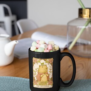 May include: Black coffee mug with a cartoon illustration of a capybara wearing a hat and a pink robe. The mug has the text "The Capybara" printed on the bottom. The mug is filled with colorful marshmallows.