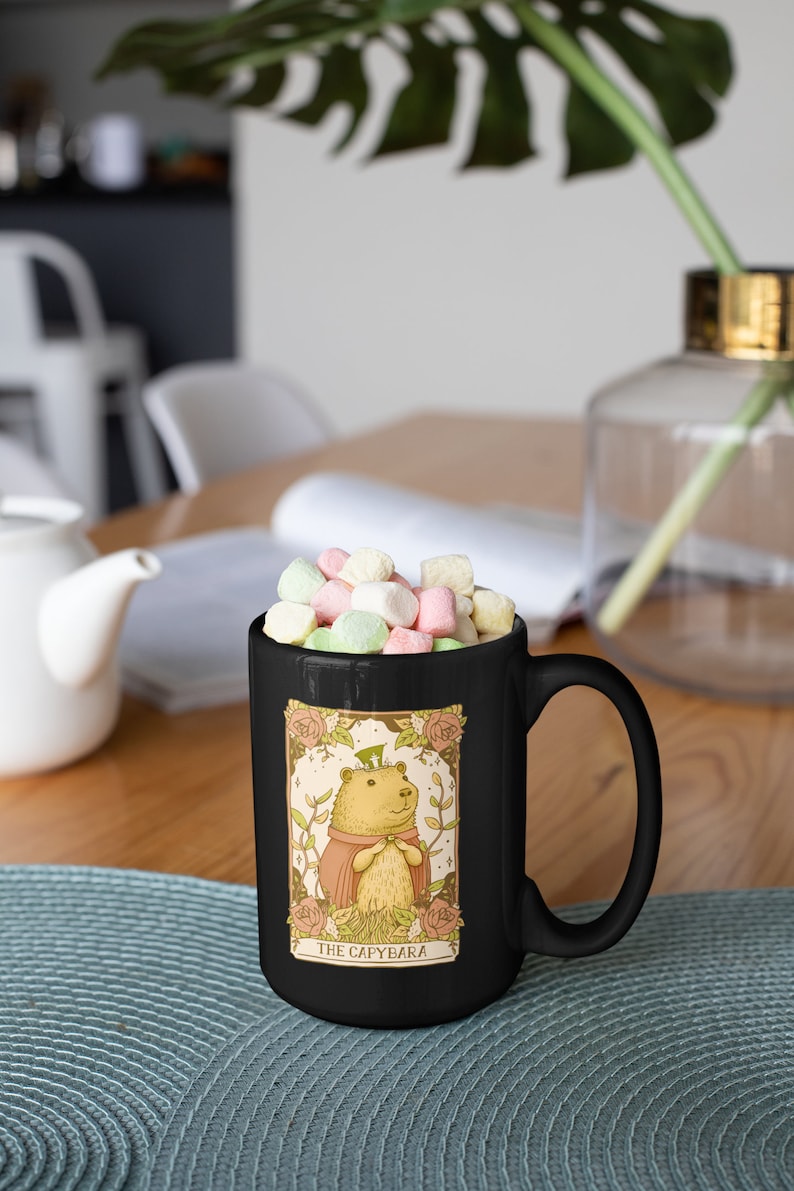 May include: Black coffee mug with a cartoon illustration of a capybara wearing a hat and a pink robe. The mug has the text "The Capybara" printed on the bottom. The mug is filled with colorful marshmallows.