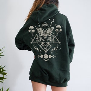 May include: A dark green hooded sweatshirt with a beige graphic print on the back. The graphic features a moth, mushrooms, moons, and stars in a geometric design.