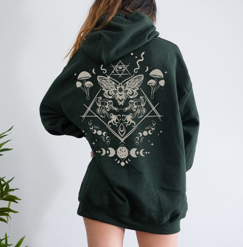 May include: A dark green hooded sweatshirt with a beige graphic print on the back. The graphic features a moth, mushrooms, moons, and stars in a geometric design.