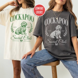 May include: Two women wearing oversized t-shirts with a vintage-style graphic of a Cockapoo dog and the text "Cockapoo Social Club Est. 1997 Santa Rosa California". The shirts are in white and charcoal grey.