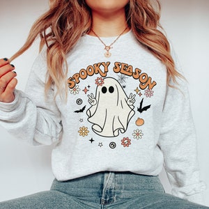 May include: A gray sweatshirt with a white ghost wearing a flower crown and the text "Spooky Season" in orange and yellow letters. The ghost is holding up two peace signs and there are flowers, stars, and bats around it.