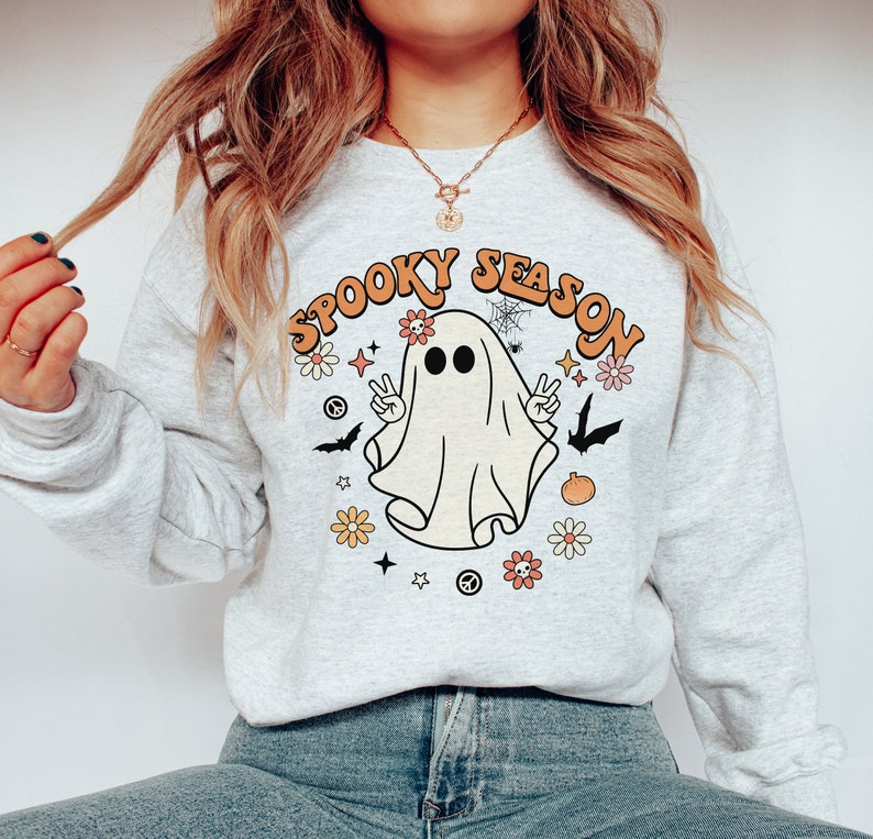 May include: A gray sweatshirt with a white ghost wearing a flower crown and the text "Spooky Season" in orange and yellow letters. The ghost is holding up two peace signs and there are flowers, stars, and bats around it.