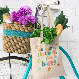 May include: A white canvas tote bag with the text "do things that make you happy" in rainbow colors and smiley faces. The bag is hanging on a blue bicycle with a wicker basket filled with flowers and groceries.