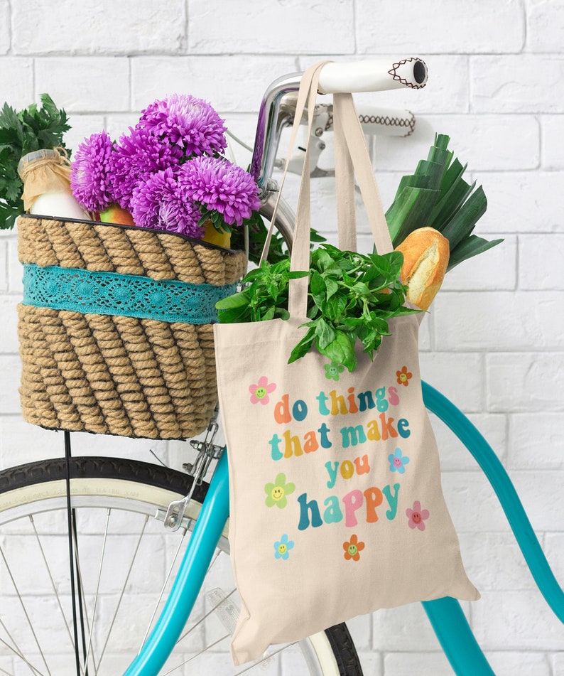 May include: A white canvas tote bag with the text "do things that make you happy" in rainbow colors and smiley faces. The bag is hanging on a blue bicycle with a wicker basket filled with flowers and groceries.