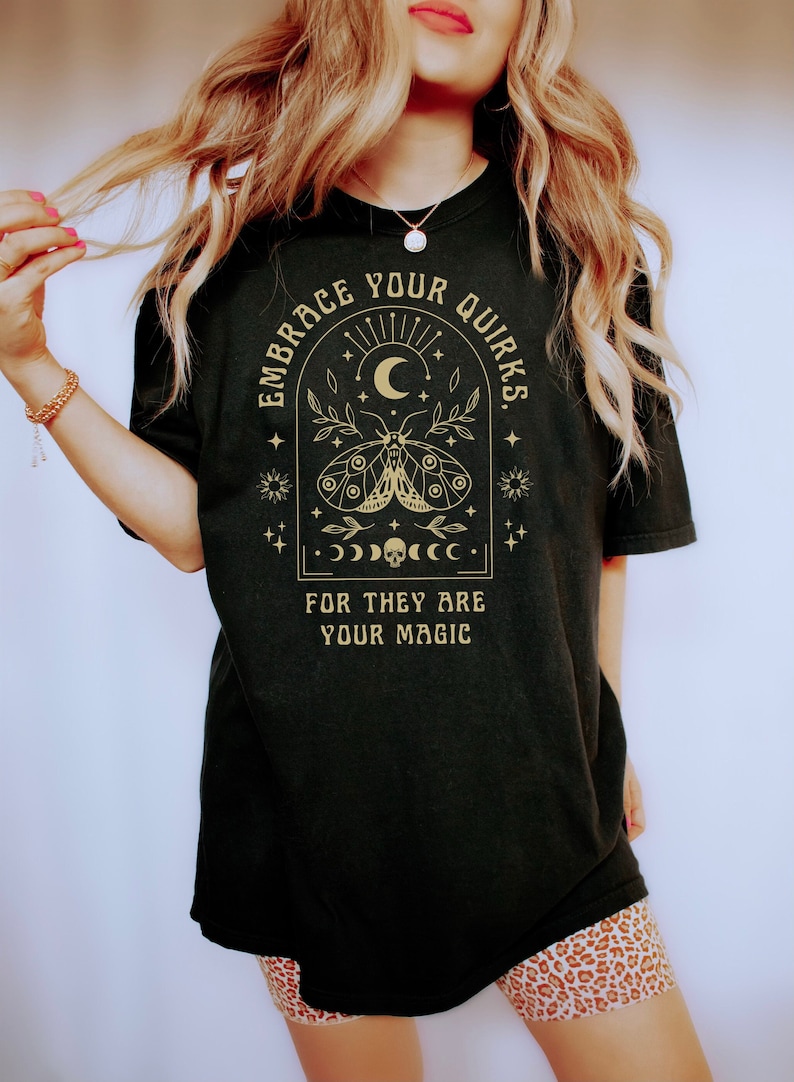 May include: Black t-shirt with a gold graphic design featuring a moth, crescent moon, stars, and the text "Embrace Your Quirks. For They Are Your Magic."