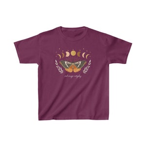 Peut inclure: Un t-shirt bordeaux avec un motif de papillon aux ailes d&eacute;ploy&eacute;es, entour&eacute; d'un croissant de lune et d'&eacute;toiles. Le texte "seek magic everyday" est imprim&eacute; sous le papillon.