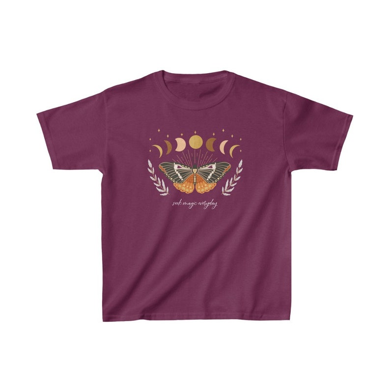Peut inclure: Un t-shirt bordeaux avec un motif de papillon aux ailes d&eacute;ploy&eacute;es, entour&eacute; d'un croissant de lune et d'&eacute;toiles. Le texte "seek magic everyday" est imprim&eacute; sous le papillon.