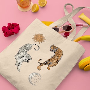 Op de afbeelding: Een beige tote bag met een witte tijger, een oranje tijger, een zon en een maanontwerp. De tas heeft twee lange hengsels.