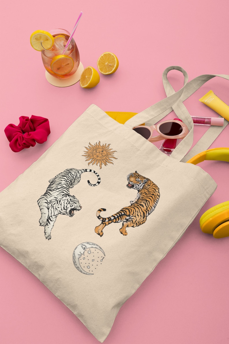 Op de afbeelding: Een beige tote bag met een witte tijger, een oranje tijger, een zon en een maanontwerp. De tas heeft twee lange hengsels.