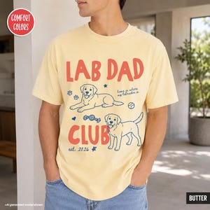 Puede incluir: Una camiseta de manga corta de color amarillo p&aacute;lido con el texto rojo LAB DAD CLUB e ilustraciones azules de dos perros labrador, una pelota y huellas. El texto dice home is where my labrador is y est. 2026. Una insignia roja dice COMFORT COLORS.