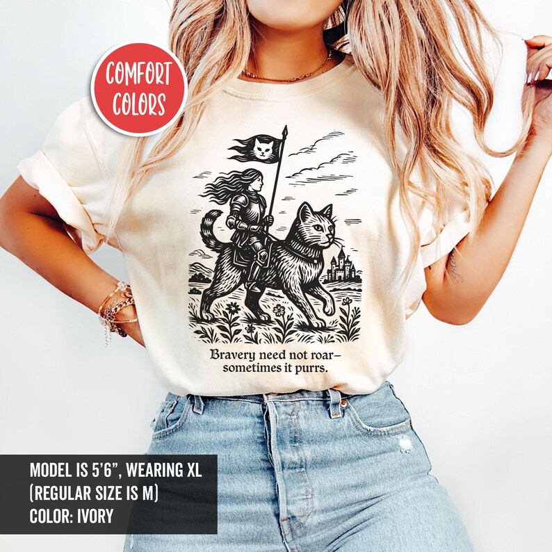 Peut inclure: Un t-shirt blanc avec une illustration en noir et blanc d'une femme chevauchant un chat. Le chat porte un collier et la femme tient un drapeau avec un chat dessus. Le texte "Bravery need not roar- sometimes it purrs." est imprim&eacute; sous l'illustration.