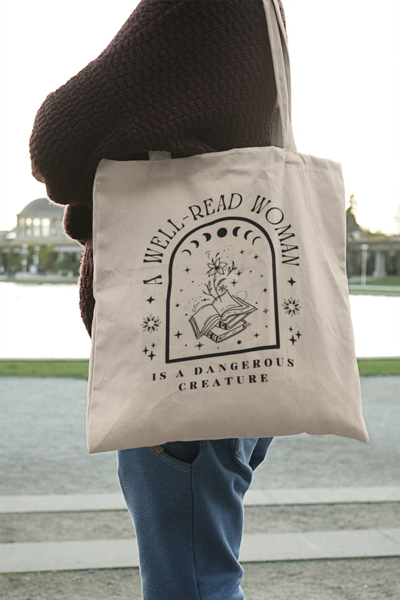 Pode incluir: Saco tote bege com o texto "A WELL-READ WOMAN IS A DANGEROUS CREATURE" em preto. O design inclui uma ilustra&ccedil;&atilde;o de livros, flores e elementos celestes. O saco &eacute; carregado por uma pessoa que usa um su&eacute;ter escuro e jeans azuis.